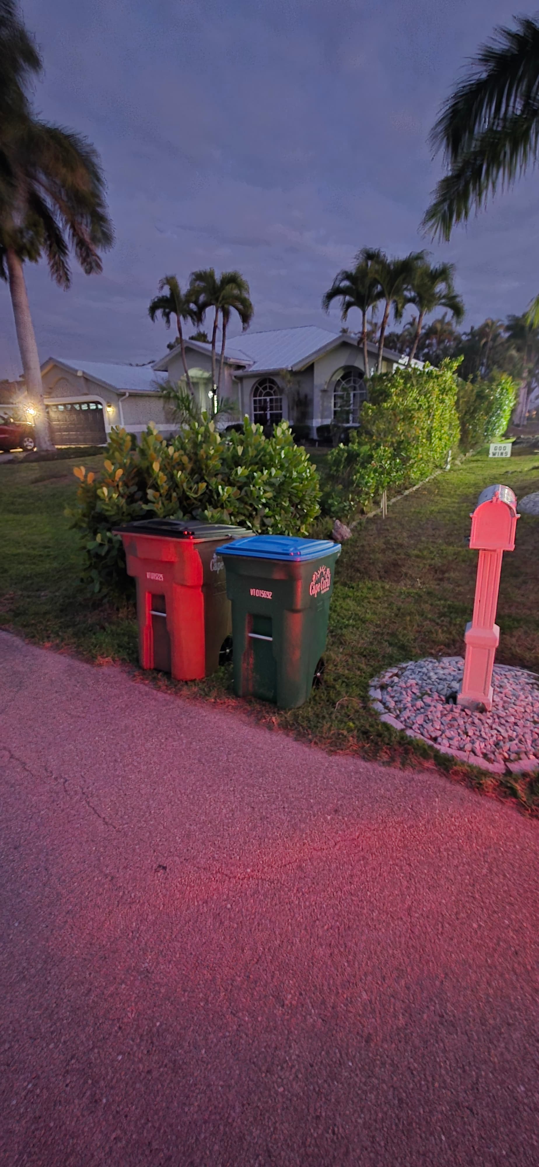Green and red trash bins on a lawn beside a driveway illuminated by red light.